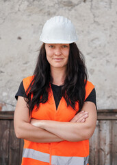 Young woman in white hard hat and orange high visibility vest, long dark hair, hands crossed, looking confident. Blurred construction site wall background