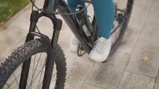 woman pedaling bicycle on pavement, hands on handlebars and feet on pedals, front wheel and fork in frame, leggings and sneakers in motion, pushing off for commute or training, momentum building,