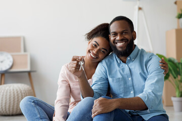 Obraz premium Happy young black couple hugging and showing keys to new apartment with cardboard boxes, looking at camera after relocation. Buying own real estate, renting apartment, moving, positive emotions
