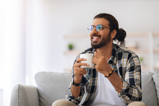 Cheerful young indian man with coffee mug looking at copy space, sitting on couch in cozy living room, happy middle-eastern guy in casual relaxing alone during weekend, drinking herbal tea