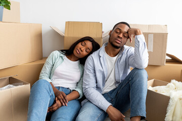Real estate and renting issues concept. Sleepy african american couple sitting among cardboard boxes, being tired on moving day. Exhausted spouses taking break in unpacking things