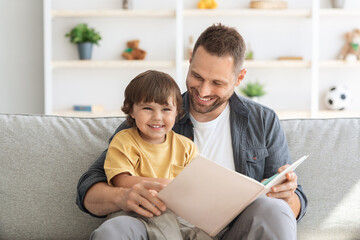 Naklejka premium Family and upbringing concept. Close up portrait of happy little boy enjoying storytelling time with his daddy, sitting in armchair with big book