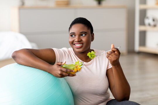 Sports and healthy nutrition for weight loss concept. Curvy black lady leaning on fitness ball, eating fresh vegetable salad at home. Plus size young woman enjoying slimming and exercising program
