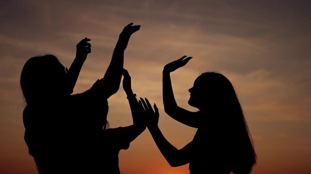 Silhouette high five at sunset, woman friend raise hand toward glowing sun in colorful sky, dark profile shape and playful gesture convey celebration connection energy during warm evening together
