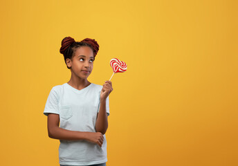 Cute african american teen girl with nice hairstyle holding candy on yellow studio background and...