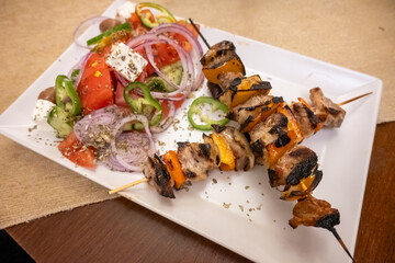 Two flavorful grilled pork and pepper skewers served alongside a vibrant Greek salad with feta, olives, and fresh vegetables on a white plate.