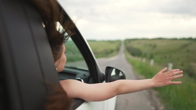 Hand reaching out car window over gravel road through green countryside with wind tousling hair of girl leaning out during travel summer drive conveying freedom and relaxed journey along rural road