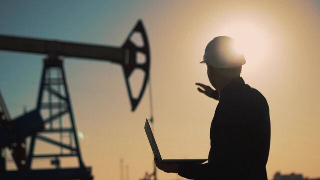 Engineer inspects oil pumpjack at sunset while holding laptop and wearing hard hat worker silhouette against glowing sky with sun behind industrial structure showing energy industry and safety