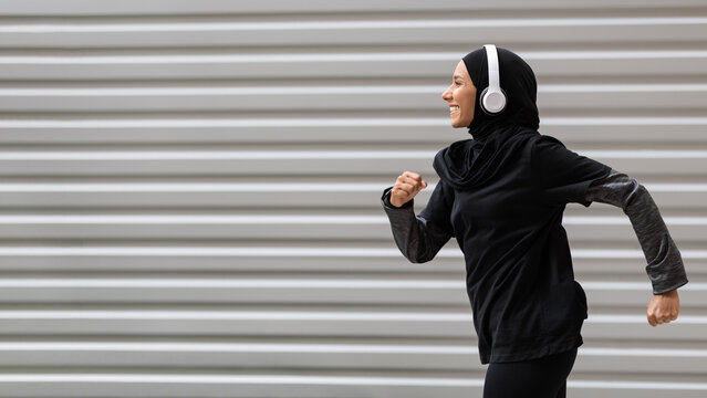 Fit muslim woman in modest sportswear and wireless headphones running outdoors, side view shot in motion of athletic islamic female runner in hijab doing sports outside, panorama with copy space