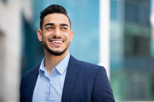 Closeup portrait of handsome young bearded arab man in suit entrepreneur or manager posing by modern business center, smiling and looking at copy space. Business opportunities concept