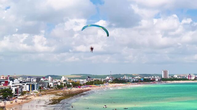 Aerial view of Jo&atilde;o Pessoa beach in Para&iacute;ba, Brazil, featuring turquoise ocean, sandy shoreline, urban skyline, and paragliding over a vibrant tropical coastal city.