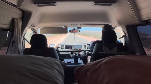 Two people are in a camper van traveling on a straight road through a desert area during the daytime.