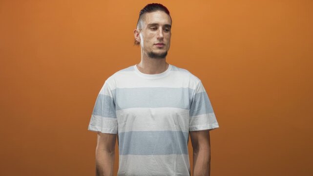 Man in striped shirt turns head to left showing profile, hands relaxed at sides against orange studio backdrop; contemplation.