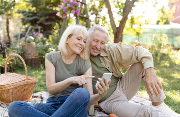 Happy senior spouses sitting on blanket during picnic in garden and using smartphone, websurfing or shopping online, resting outdoors on warm spring day