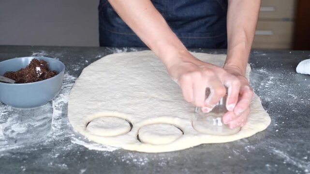 Person prepares cookie dough on a kitchen counter. Round pieces of dough are being cut using a cutter. Flour is spread around for easy handling. Ingredients are nearby. 