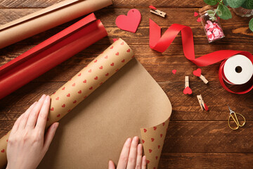 Hands unrolling heart-patterned craft paper on wooden table. Gift wrapping for Valentine's Day or wedding with red ribbon, scissors, and decor.
