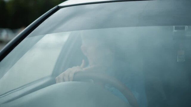 white male driver behind foggy windshield gripping steering wheel, pensive expression, blurred reflection across glass, urban road visible through mist, interior closeup showing dashboard and hands,