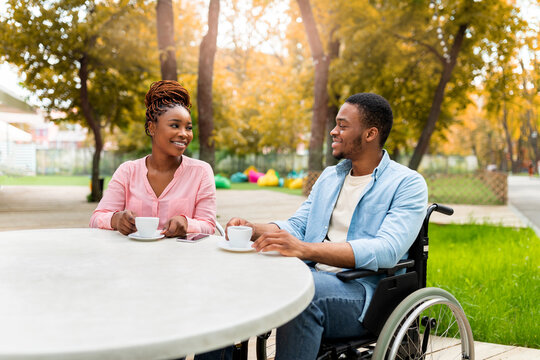 Disabled millennial black guy in wheelchair having date with lovely woman in outdoor cafe, drinking coffee, enjoying friendly talk at autumn park. Impaired couple having romantic times outside