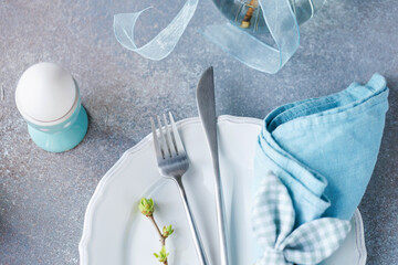 Spring table setting with white plate, cutlery, egg holder, and blue napkin on stone background