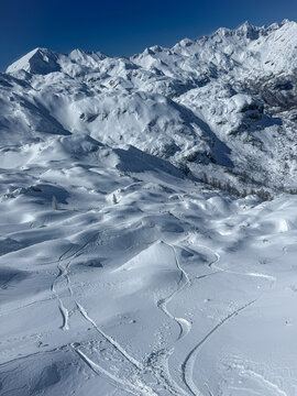 Rolling alpine ridges and bowls blanketed in fresh snow descend toward valley hidden beneath white peaks. Winding freeride ski and snowboard tracks form a pattern across vast winter mountain landscape