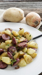 Dish of fried potatoes and sausages served on a plate on wooden table with herbs and utensils
