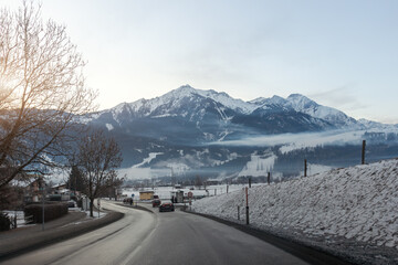 Winter alpine road leading through quiet village snowy mountains misty valley, forested slopes pale sky Kitzsteinhorn peak background. Scenic countryside route shows cold weather travel tranquility