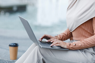 Work in city, freelance, independent contractor, digital nomad, remote job. Millennial middle eastern female in hijab with cup of coffee takeaway typing on laptop, sitting on city stairs, cropped