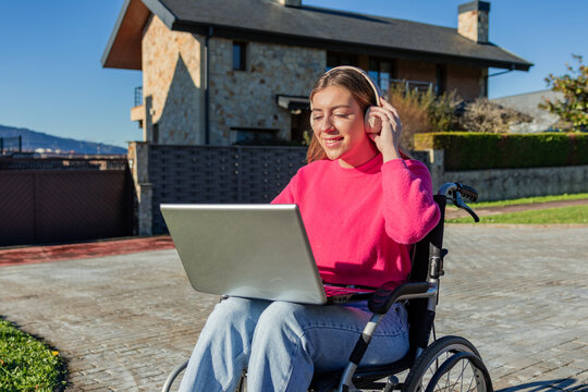 Young woman in wheelchair using laptop and headphones outdoors