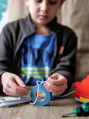 Child Holding Exposed Electrical Wires with Insulation Tape - Young Boy Learning Electrical Repair Skills at a Wooden Table
