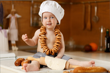 Cute baby in chef hat playing with dough and bagels on a kitchen counter