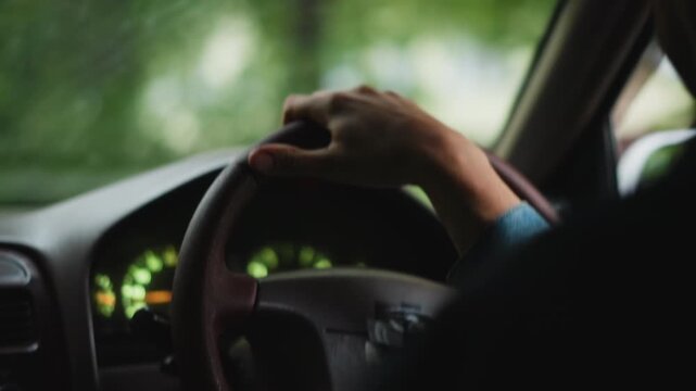 Closeup hands on steering wheel, steady grip on leather wheel, illuminated speedometer, denim sleeve visible, blurred green trees outside window, calm commuting mood and focused control