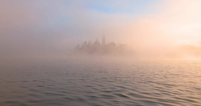 Die Kirche auf der Insel im See Bled im Morgengrauen eingerahmt von gold gef&auml;rbtem Nebel im warmen Licht der goldenen Stunde vor bewaldetem Ufer und ruhigem Wasser