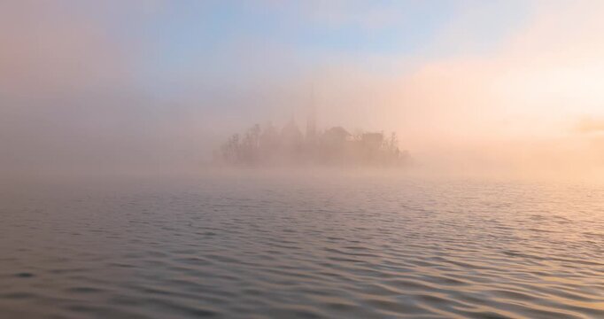 Die Kirche auf der Insel im See Bled im Morgengrauen eingerahmt von gold gef&auml;rbtem Nebel im warmen Licht der goldenen Stunde vor bewaldetem Ufer und ruhigem Wasser