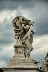 Obraz premium Baroque marble statue of winged angel figure embracing small child on ornate pedestal against dark stormy clouds at the Vittoriano monument