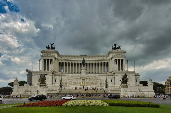 View of the white marble Vittoriano monument (Altare della Patria) with bronze quadrigas and Italian flags under dramatic stormy summer sky with tourists