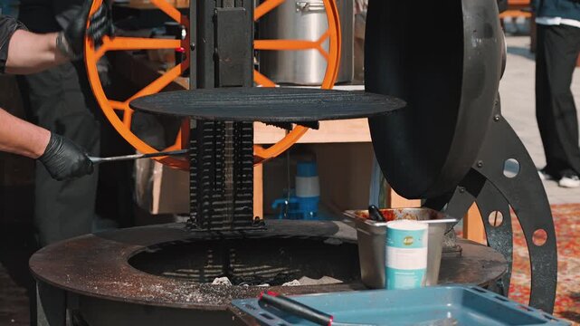 A chef is seen adjusting a large outdoor grill, showcasing a metallic circular grill plate. The vibrant orange wheel contrasts with the dark metal of the grill and surrounding equipment