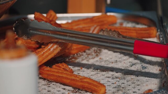 Crispy churros, dusted with sugar, are laid out on a tray, ready to be served. Stainless steel tongs lie nearby. Close-up