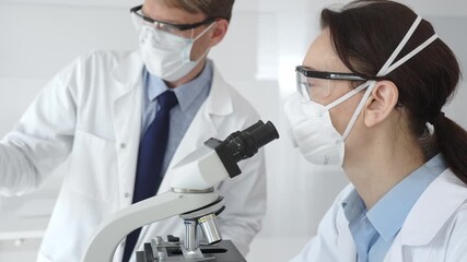 Two scientists in lab coats and safety gear conducting research, one observing through a microscope and the other holding a test tube with turquoise liquid. Medicine and science concept - Powered by Adobe