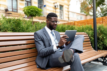 Young black businessman reading on pad while having lunch break, sitting on bench near office center, copy space. Modern using digital tablet, websurfing or checking social media