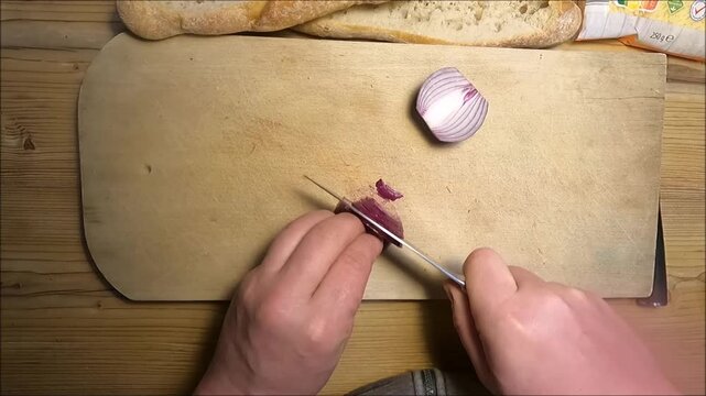 Draufsicht auf H&auml;nde beim Schneiden und Sch&auml;len einer Zwiebel auf einem Holzschneidebrett. Alltagsszene aus der K&uuml;che, die Kochen, Vorbereitung und Hausmannskost zeigt.