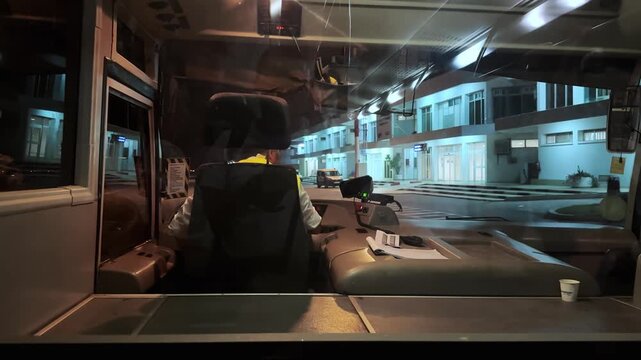 View of transfer bus driving at night showing the driver's seat and dashboard, with airport visible through the front window