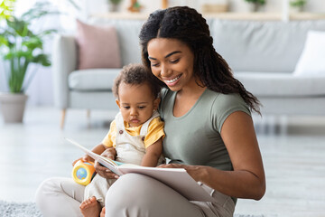 Naklejka premium Caring African American Mommy Reading Book For Her Adorable Toddler Son At Home, Young Black Woman Bonding With Cute Little Infant Baby, Enjoying Spending Time Together, Closeup Shot With Copy Space