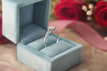 Elegant diamond engagement ring displayed in a soft blue velvet box, surrounded by romantic red roses and delicate ribbon, symbolizing love and commitment