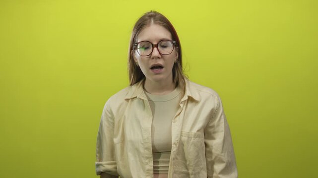 Young woman with glasses shows open mouth gesture and bare midriff in green studio setting; surprise.