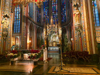 The richly decorated interior of De Krijtberg Church in Amsterdam, featuring colorful neo-Gothic details, stained glass windows, gilded statues and an ornate altar glowing with light