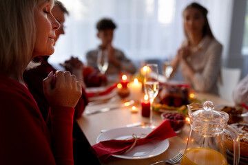 Elderly lady and her extended family praying, expressing thanks to God, sitting at table with festive meal at home, copy space. Senior woman with her nears celebrating Christmas or Thanksgiving