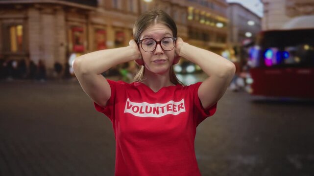 Blonde volunteer woman covers ears with hands on busy city street at night amid neon lights; stress noise overload tension.