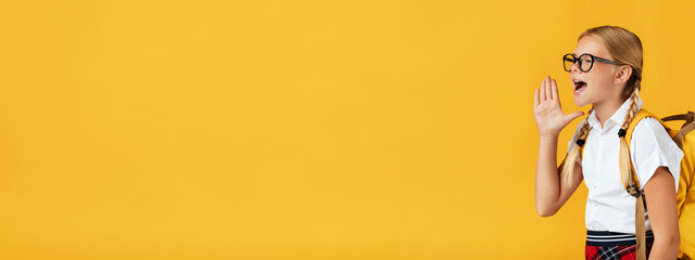Cheerful pretty teenage blonde girl in glasses shouts at free space, isolated on yellow background, panorama. Modern ad, schooling offer, education concept. Back to school, kid learning and knowledge