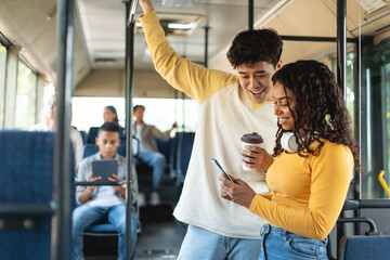 Public Transportation. Portrait of happy smiling black young lady and Asian guy taking bus together, standing using mobile phone sharing media, enjoying ride on public vehicle, drink coffee
