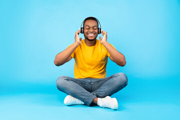 Black Guy Listening To Music With Eyes Closed Wearing Wireless Headphones Sitting In Lotus Position Over Blue Studio Background. Musical Application For Online Meditation. Relaxation Concept